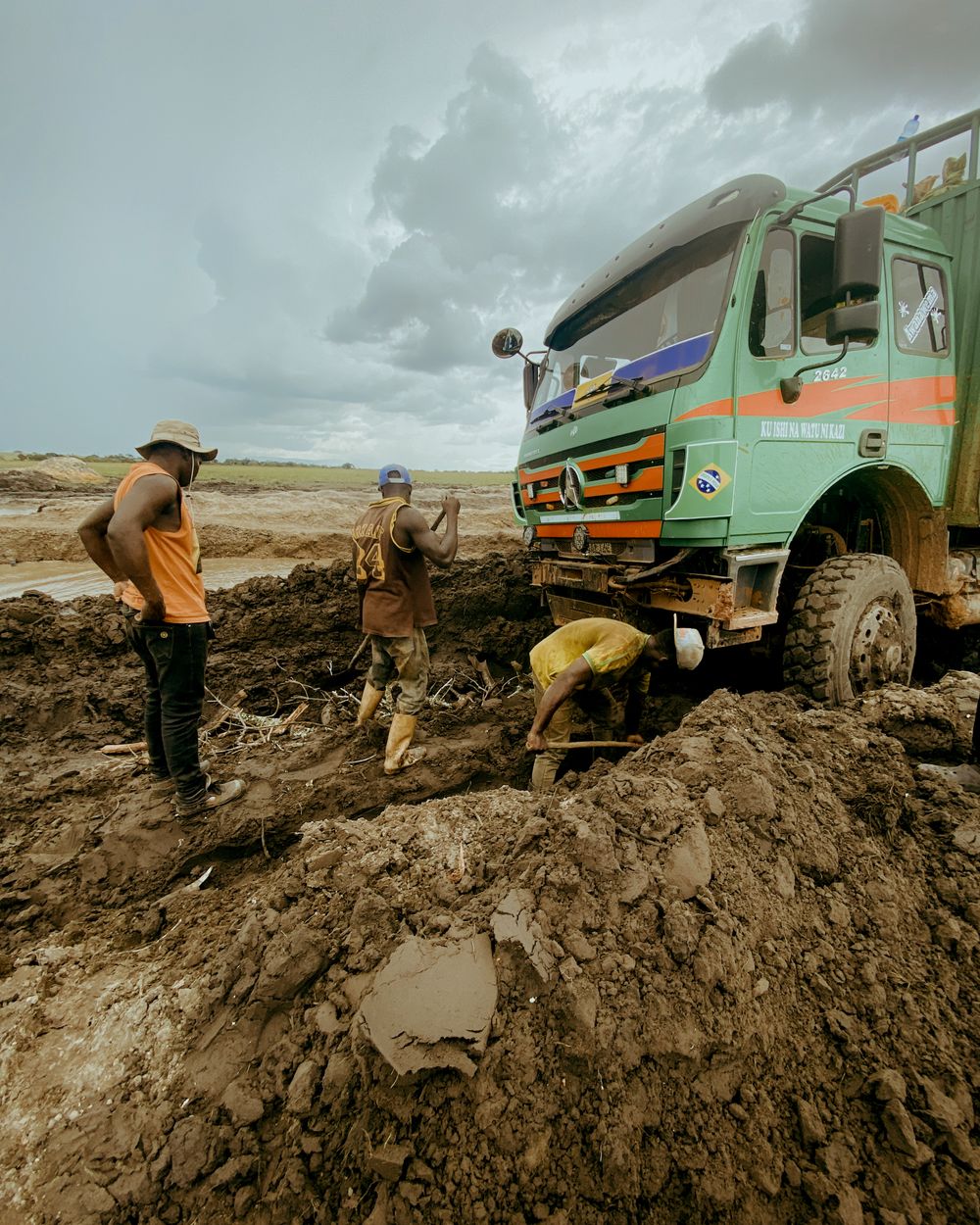 Three men dig a loaded lorry out of deep mud on an open road under a heavy storm sky